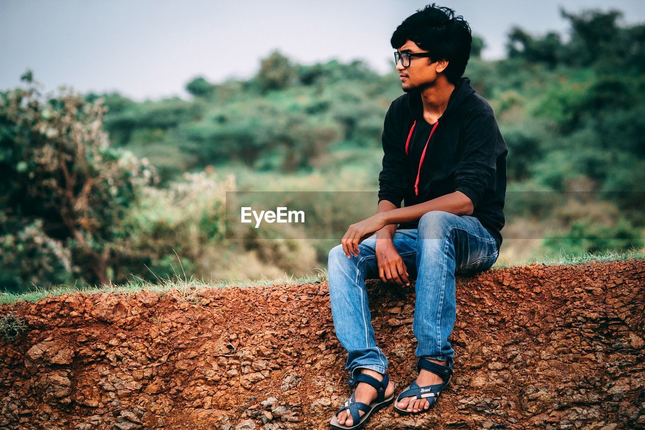 Young man looking away while sitting on field against trees