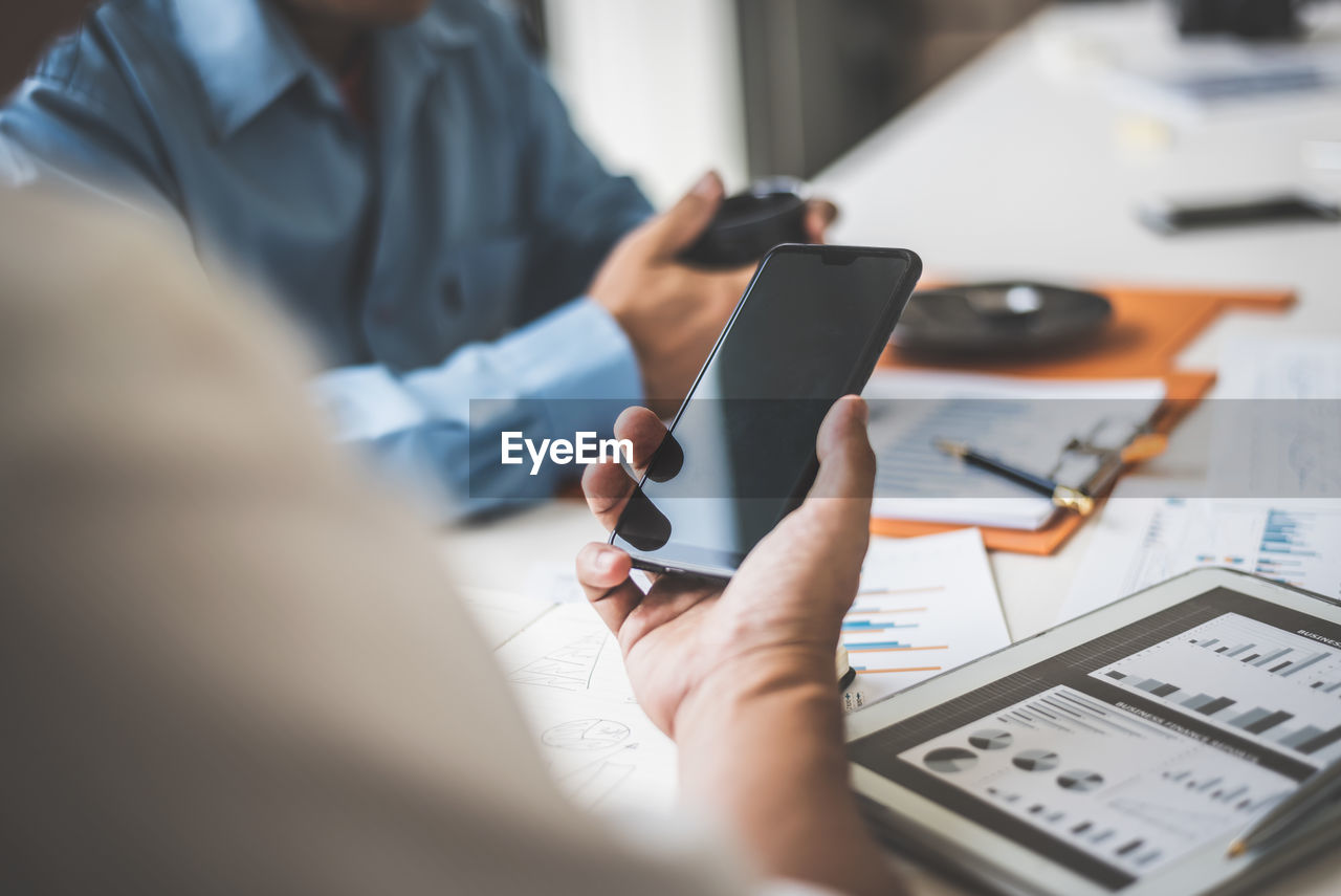 Cropped hand of businessman using phone with male coworker at desk in office