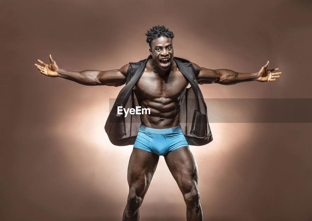 Portrait of shirtless athlete with arms outstretched standing against brown background