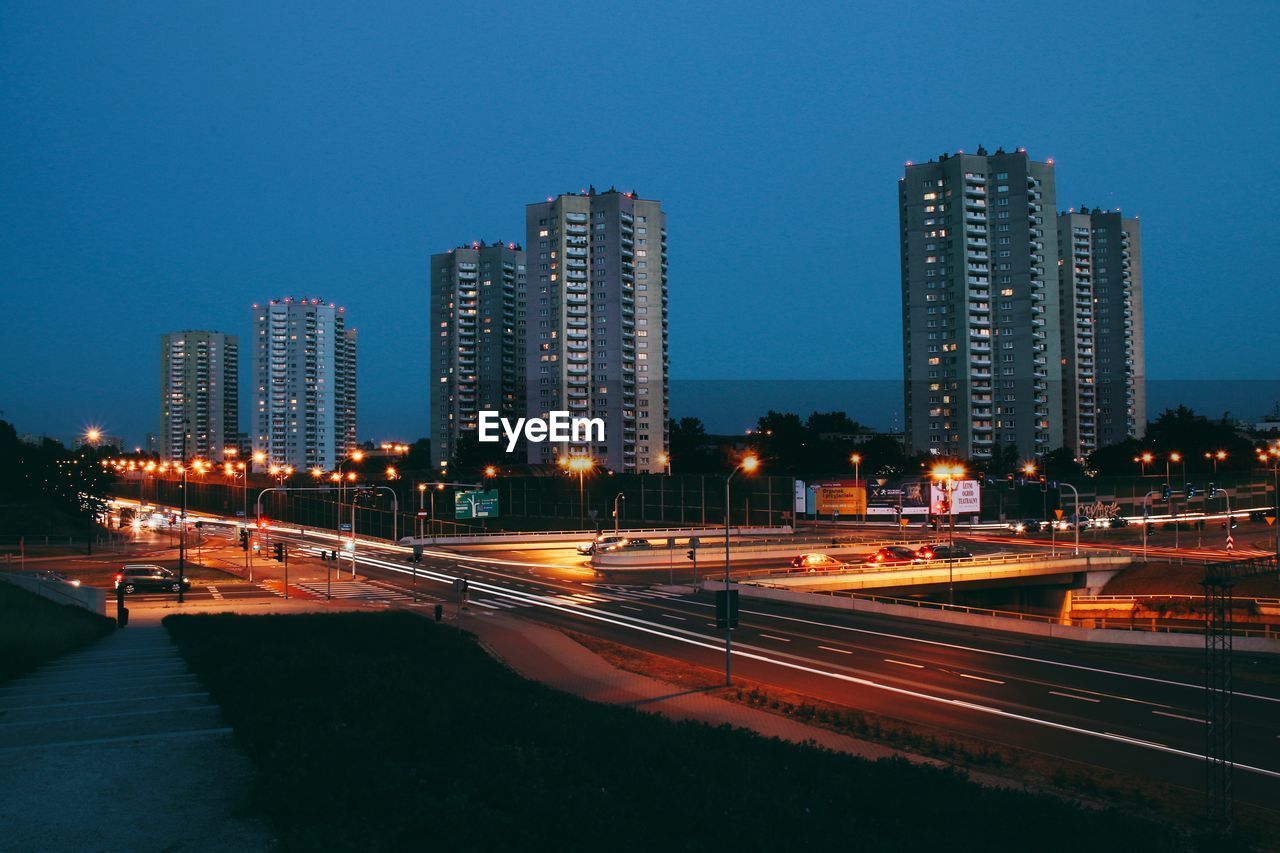 LIGHT TRAILS ON ROAD BY ILLUMINATED BUILDINGS AGAINST SKY