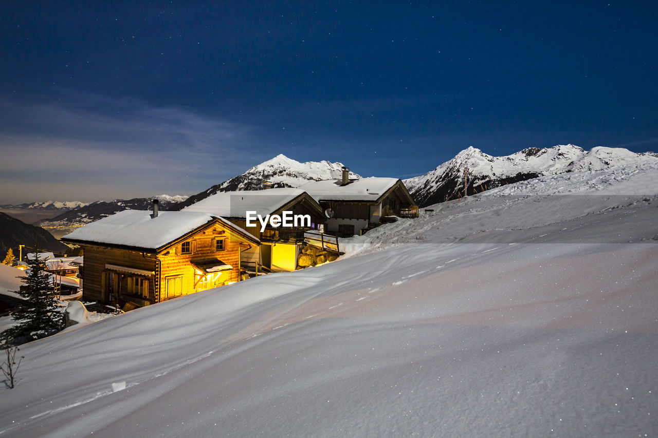 Chalets at snow covered mountain against blue sky at night during winter