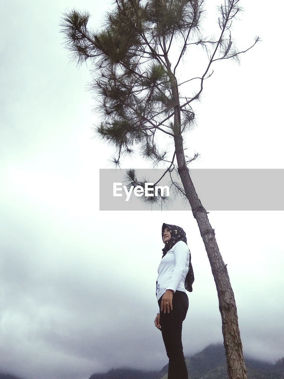 SIDE VIEW OF YOUNG MAN STANDING AGAINST TREE