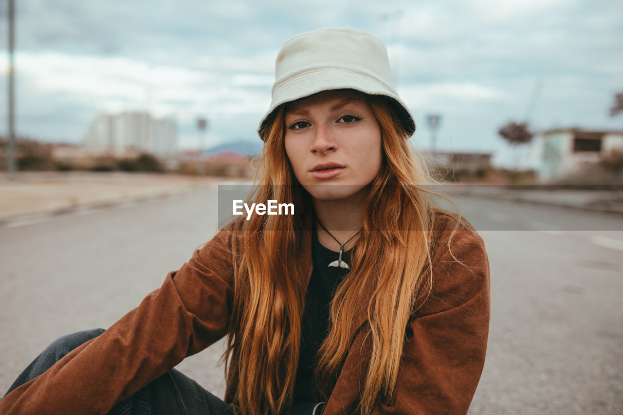 Confident young female with long ginger hair sitting on street on cloudy day and looking at camera