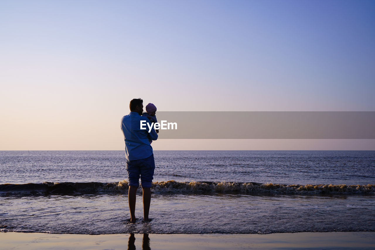 Rear view of man with toddler standing at beach against sky during sunset