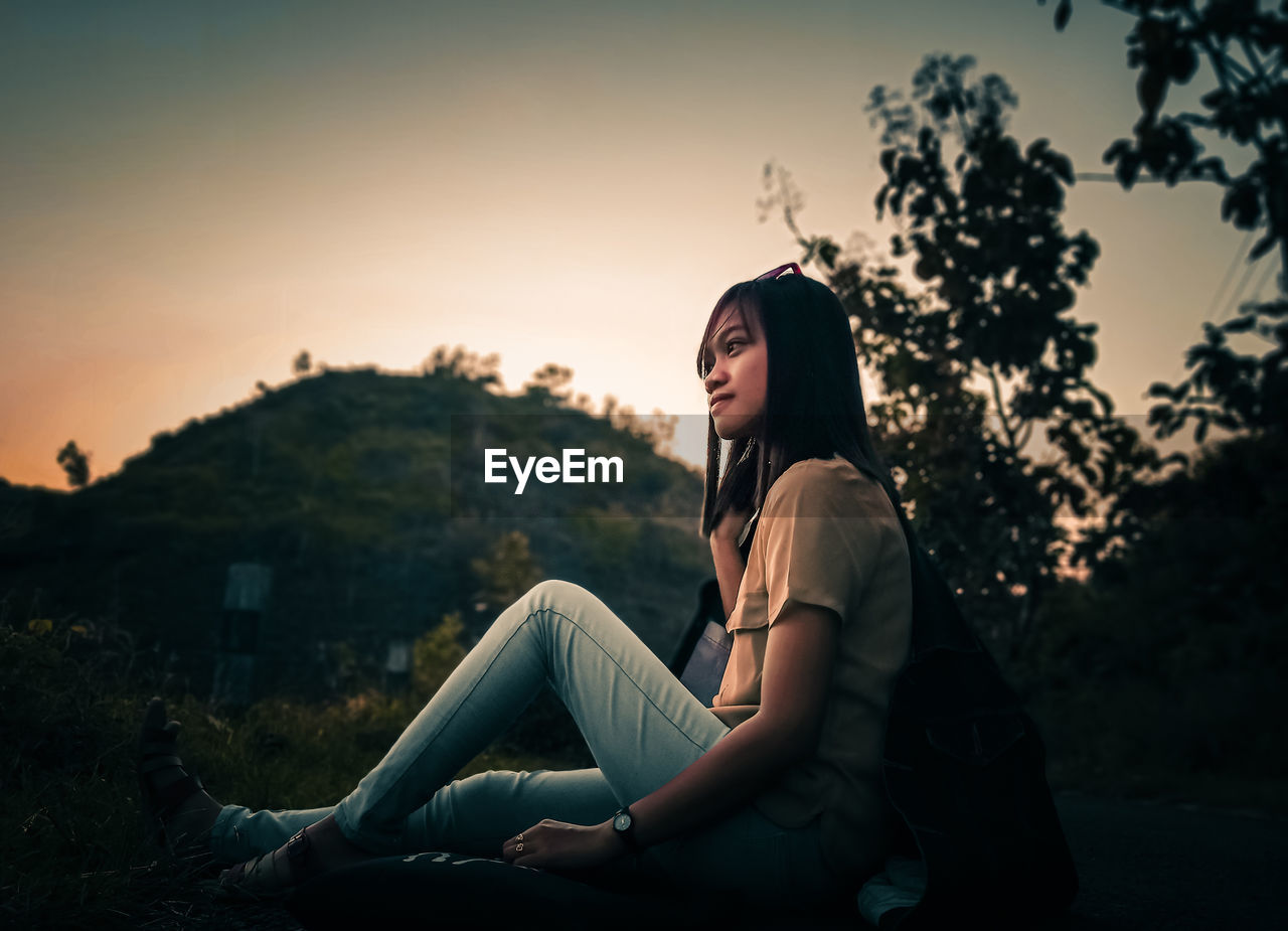 Side view of woman sitting against sky during sunset