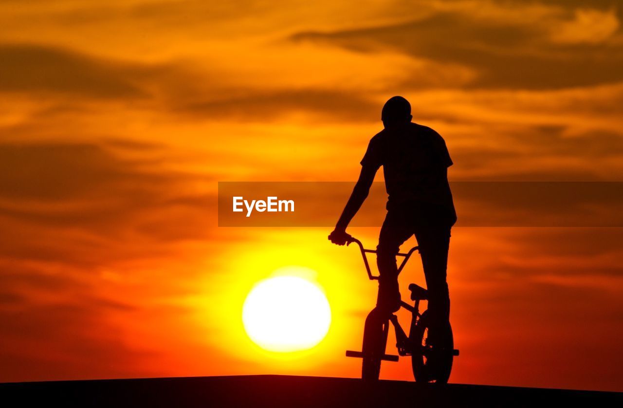 Silhouette man riding bicycle against orange sky