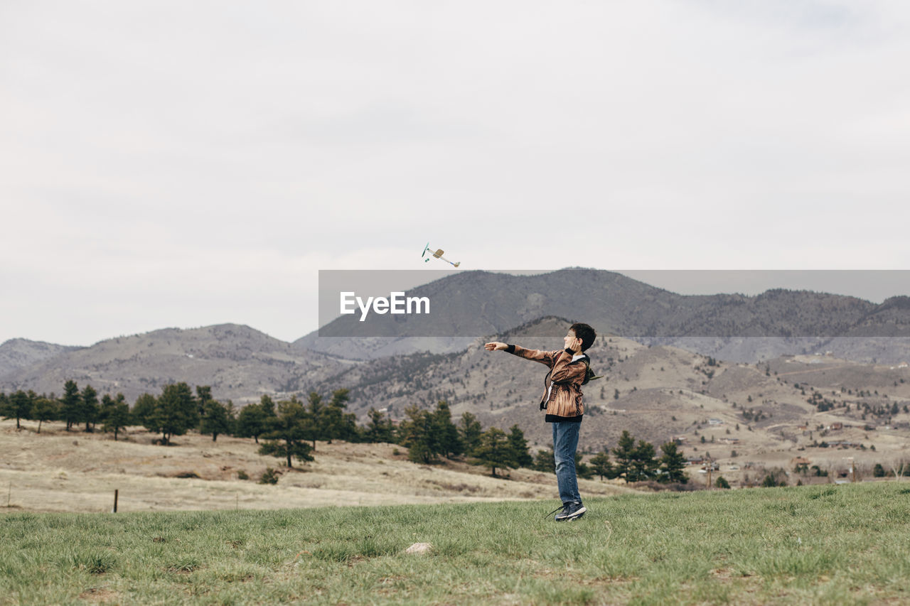 Side view of boy flying toy airplane while standing on grassy field against sky