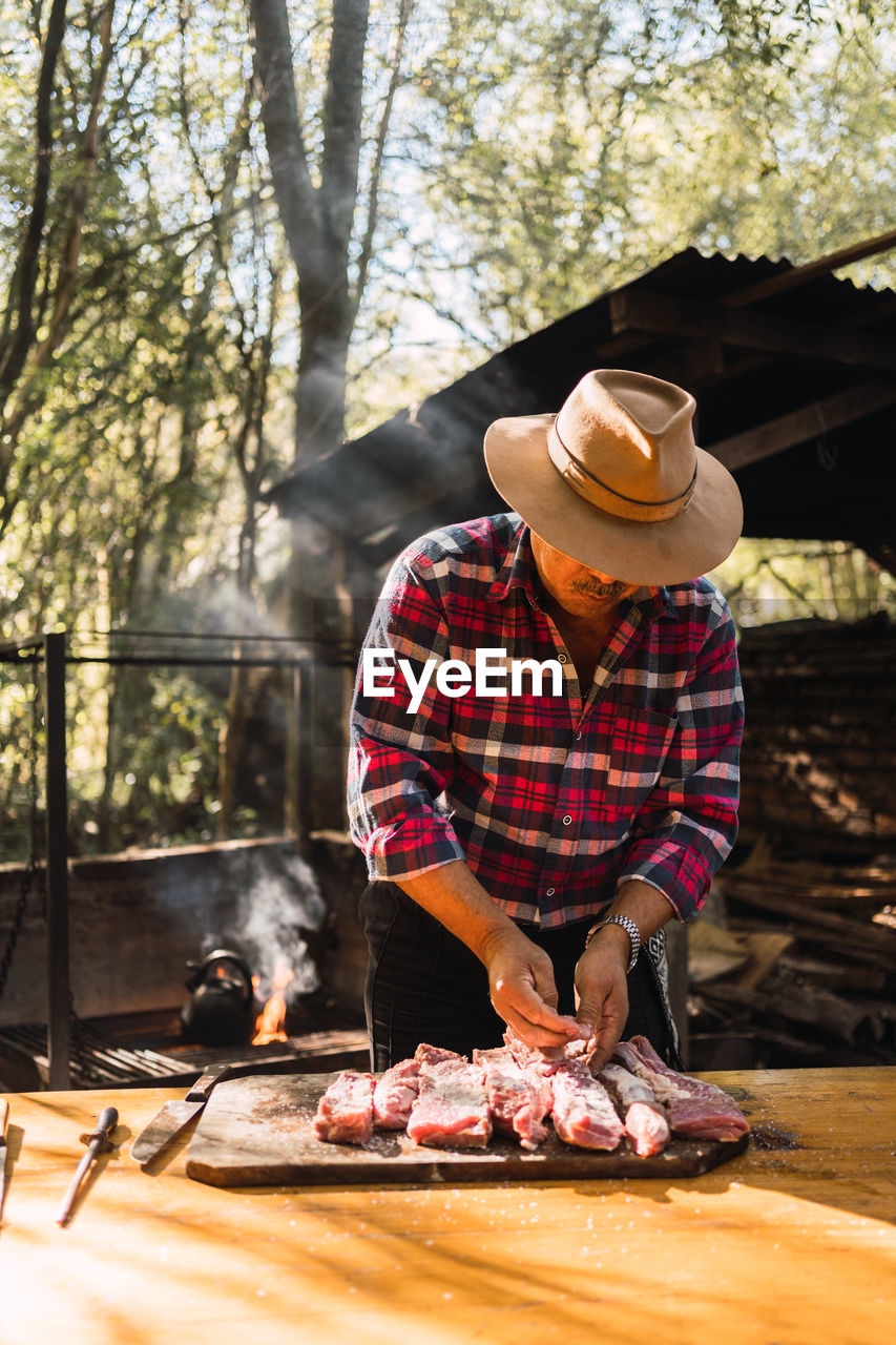 Adult argentinian male with mustache wearing beige hunter hat and checkered shirt salting meat on cutting board while preparing grill in picnic in nature