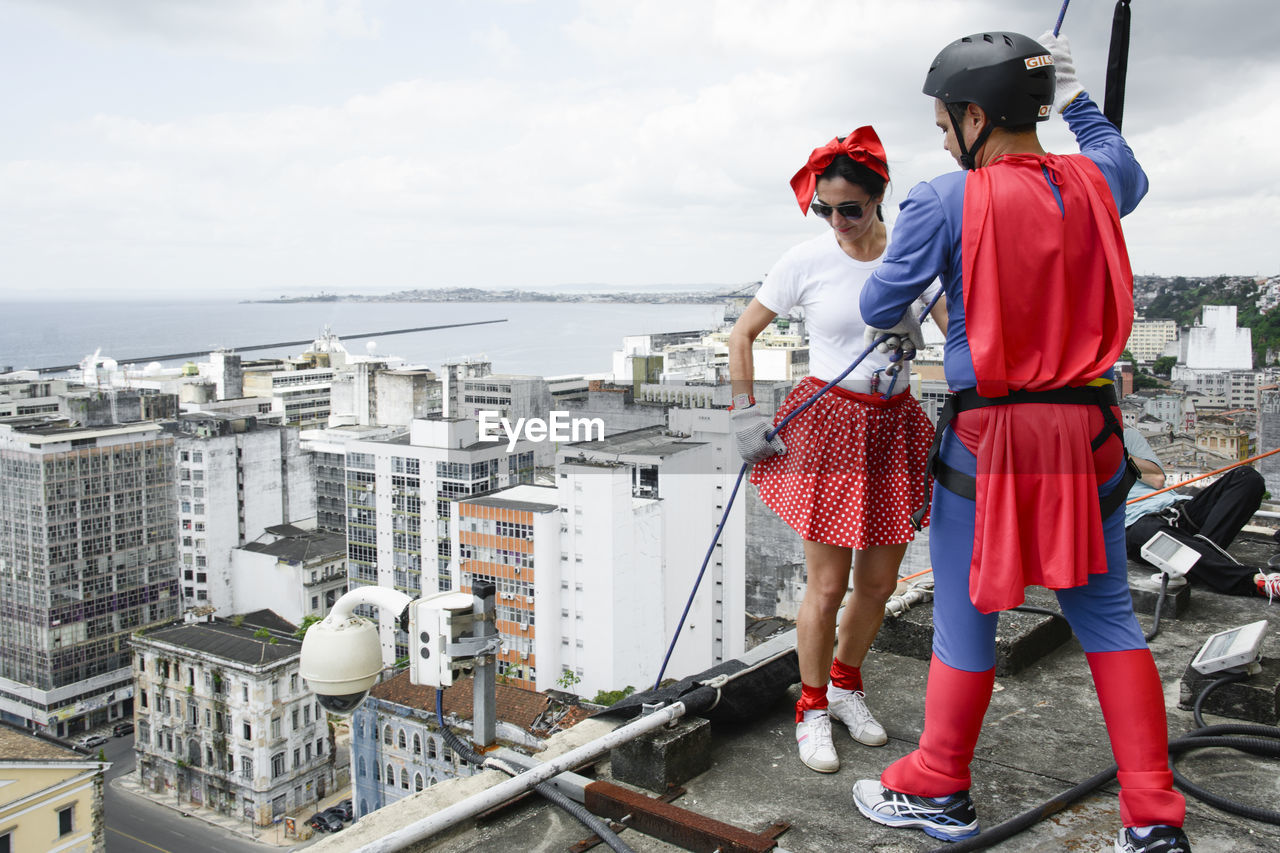 Woman wearing hero costume being prepared by a man to descend a tall rappel building. 