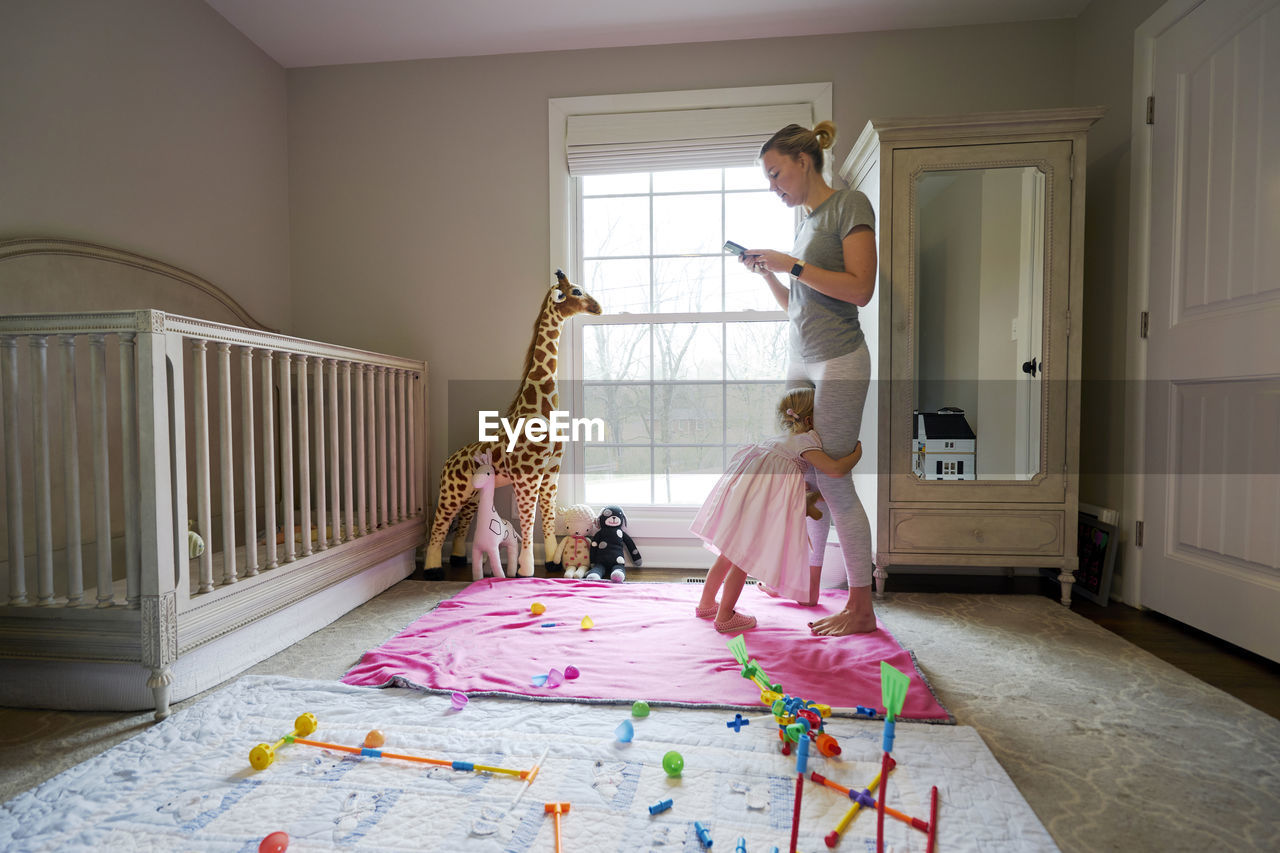 A mom looks at her phone as her toddler daughter hugs her legs