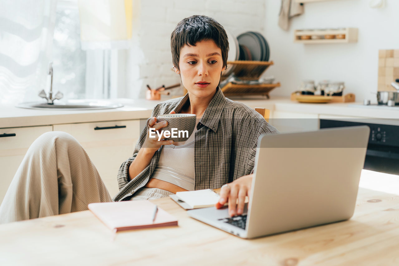 Young woman sitting at home, drinking coffee and working on laptop.