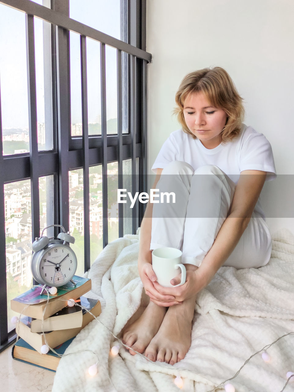 Attractive young woman with the stack of books and alarm clock in the side sitting on the windowsill