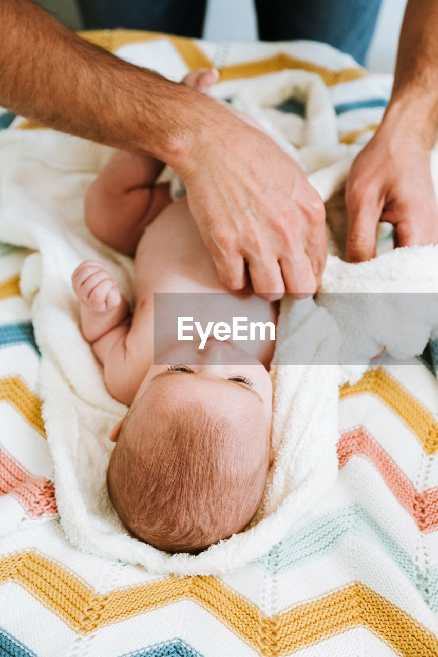 From above unrecognizable man dressing relaxed infant in soft white bathrobe while kid lying on colorful knitted plaid on bed at home