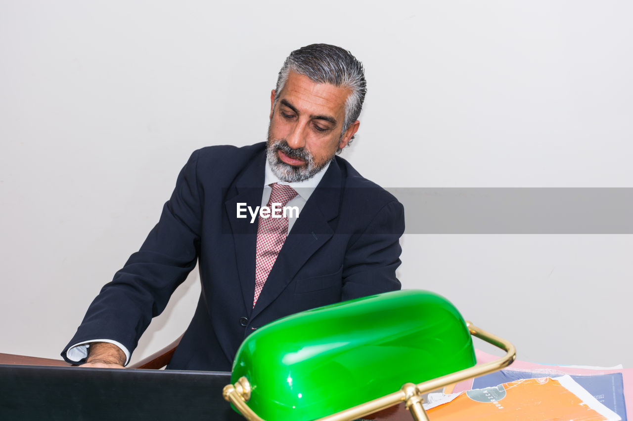 Mature businessman using laptop on desk against wall at office 