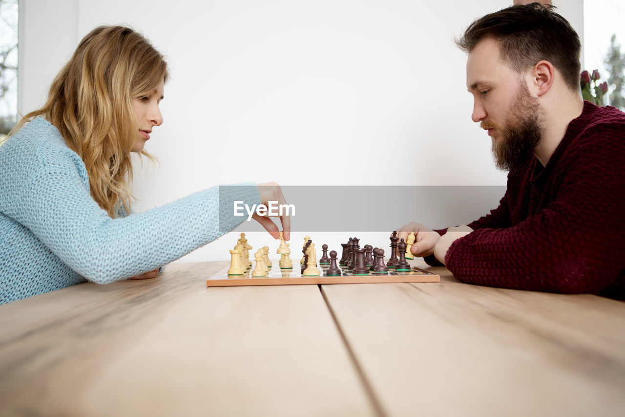 Side view of young man and woman playing chess while sitting on table at home
