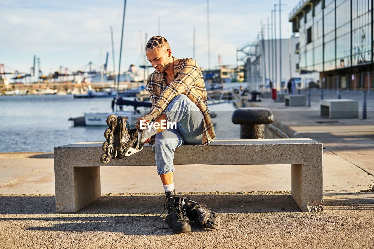 Full body of eccentric male sitting on bench and putting on roller skates on waterfront for riding at spare time