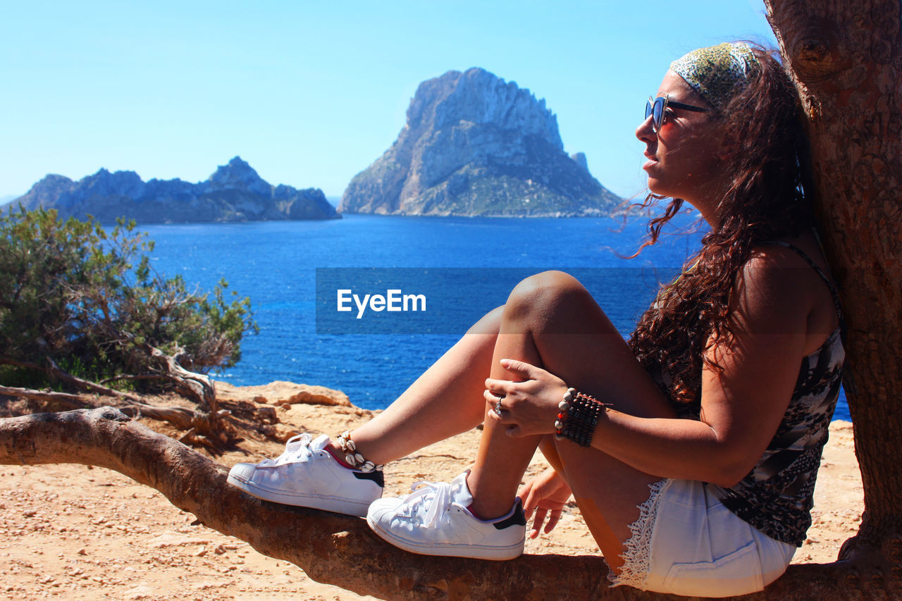 SIDE VIEW OF YOUNG WOMAN SITTING ON SEA AGAINST MOUNTAIN