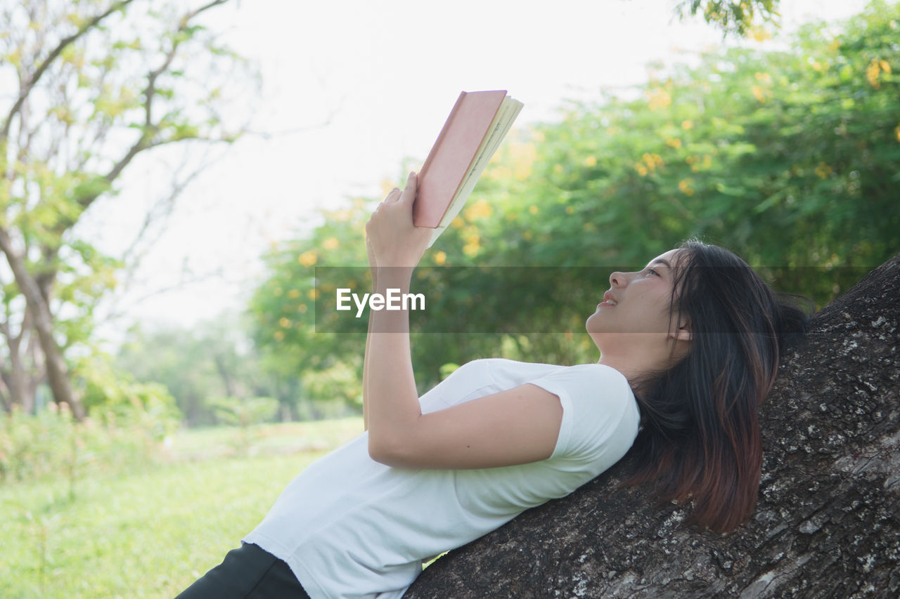 SIDE VIEW OF YOUNG WOMAN LOOKING UP WHILE STANDING AGAINST TREES