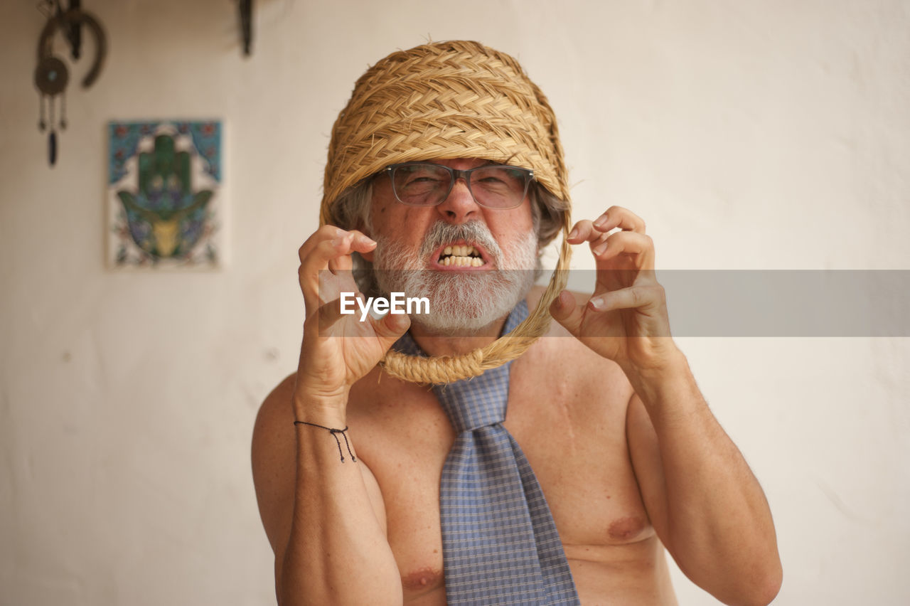 Portrait of angry senior man with basket on head standing against wall at home