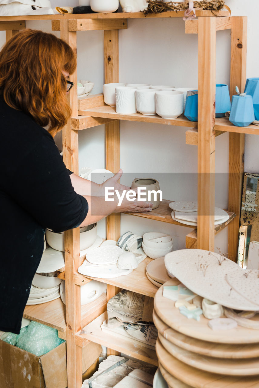 Side view of unrecognizable woman in casual black outfit standing near shelves with various pots and bowls with plates and different pottery while putting small clay vase in light studio