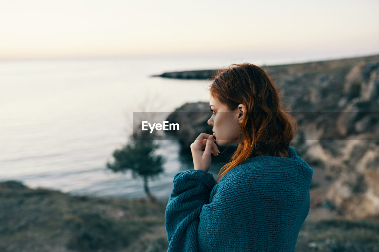 YOUNG WOMAN STANDING BY SEA AGAINST SKY DURING SUNSET