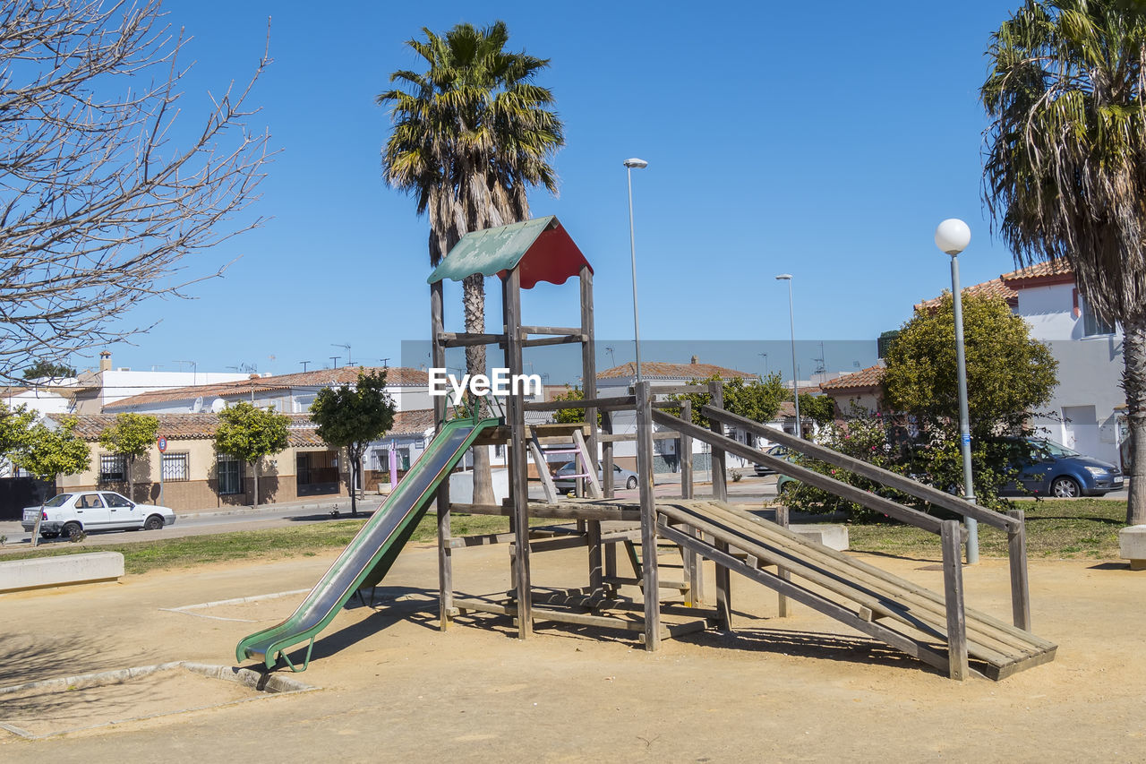 city, public space, playground, tree, outdoor play equipment, sky, nature, recreation, park, walkway, architecture, plant, palm tree, sunny, day, clear sky, tropical climate, built structure, blue, vacation, sunlight, no people, slide, outdoors, park - man made space, building exterior, playground slide