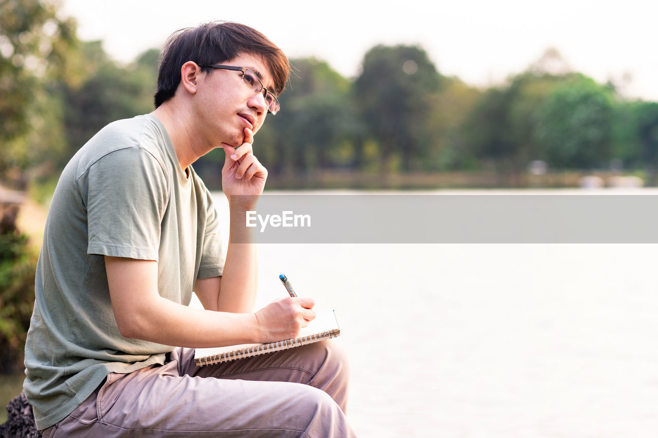 Young man looking away while sitting with book and pen by lake