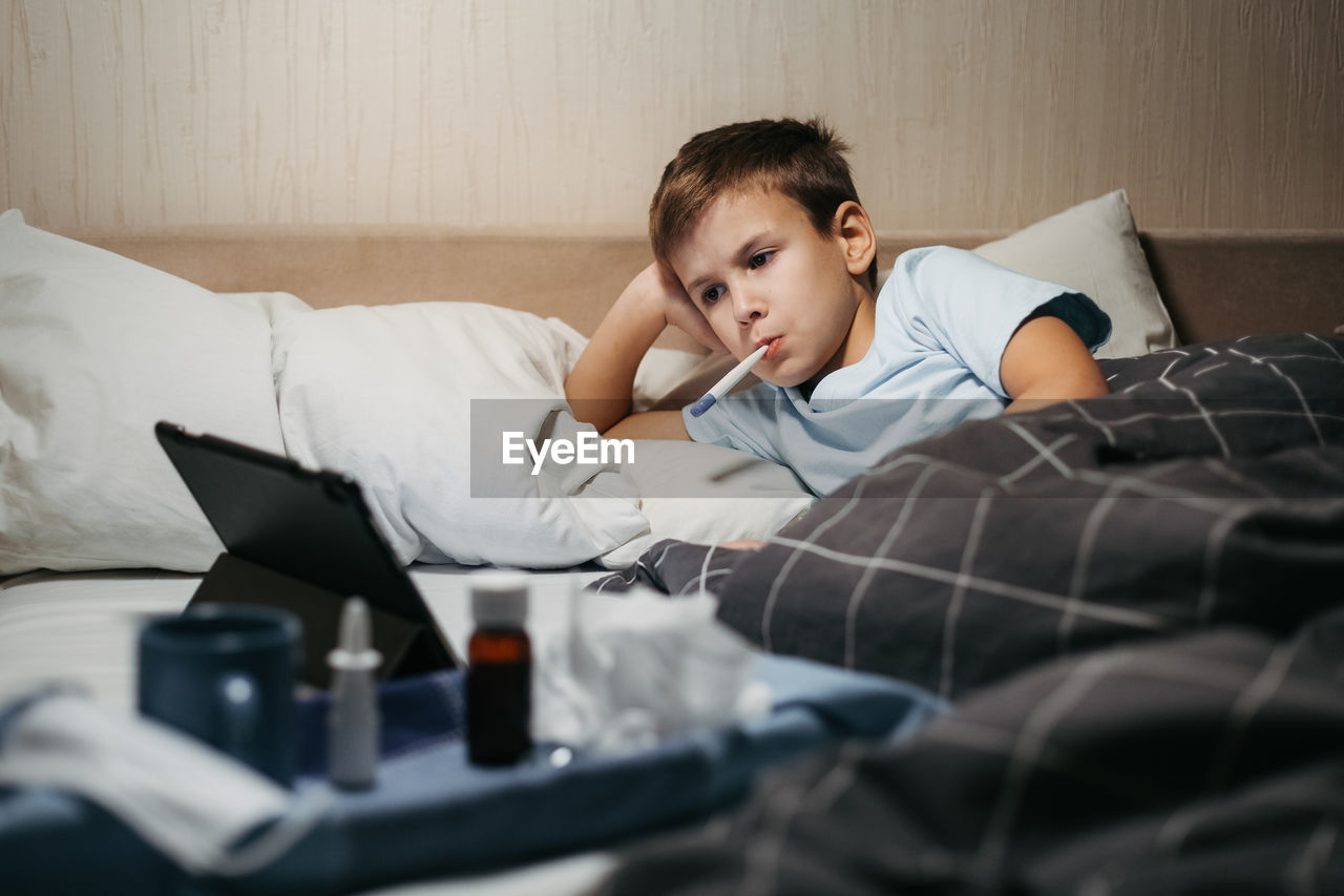 Sad child laying in bed with thermometer in his mouth and look at tablet.