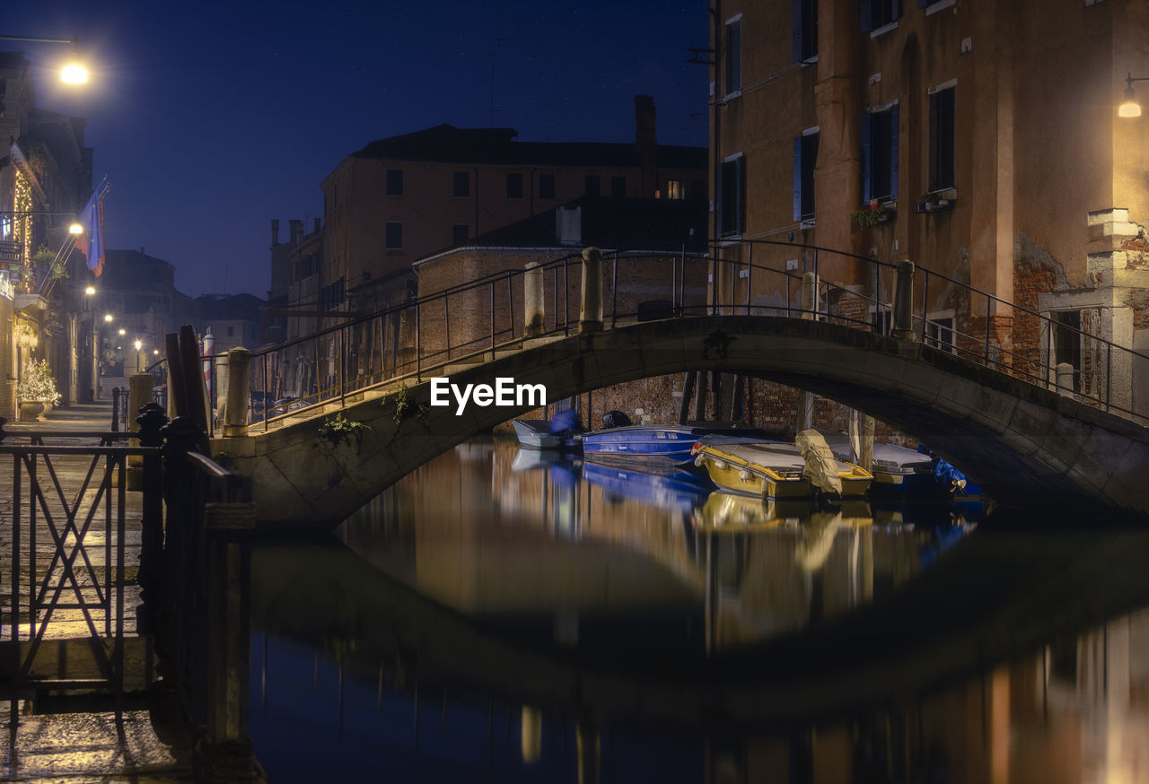 BRIDGE OVER CANAL AMIDST BUILDINGS AT NIGHT