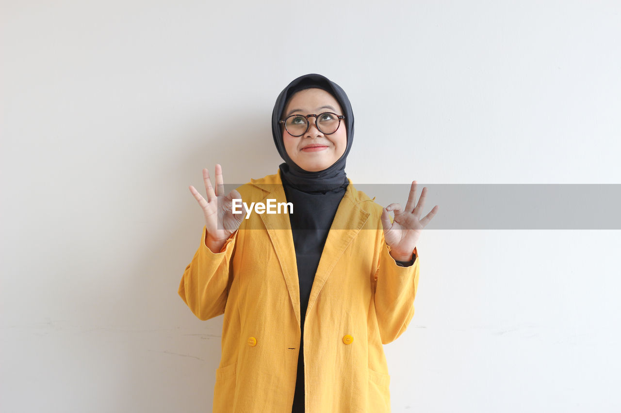 one person, yellow, clothing, studio shot, adult, portrait, indoors, front view, standing, young adult, waist up, eyeglasses, finger, hand, copy space, white background, glasses, looking at camera, emotion, person, smiling, gesturing, outerwear, human face, costume, happiness