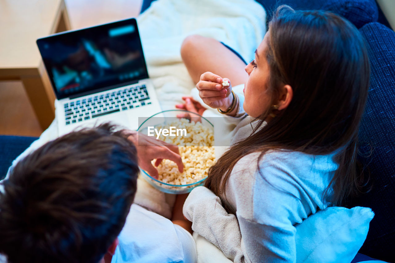 Cheerful young man and woman in casual wear eating popcorn and watching film on laptop while resting together on cozy sofa at home
