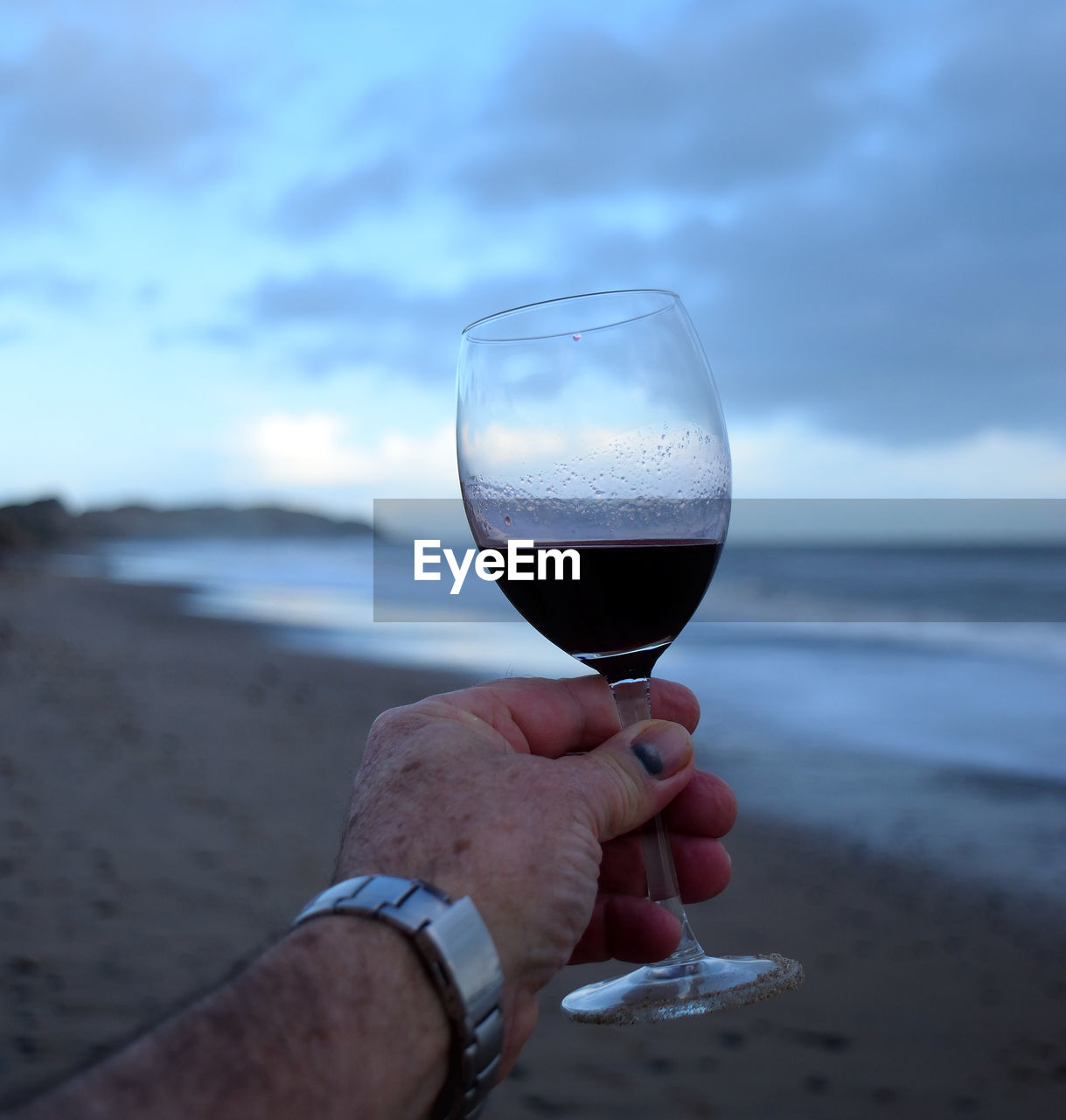 CLOSE-UP OF HAND HOLDING WINEGLASS AGAINST BEACH