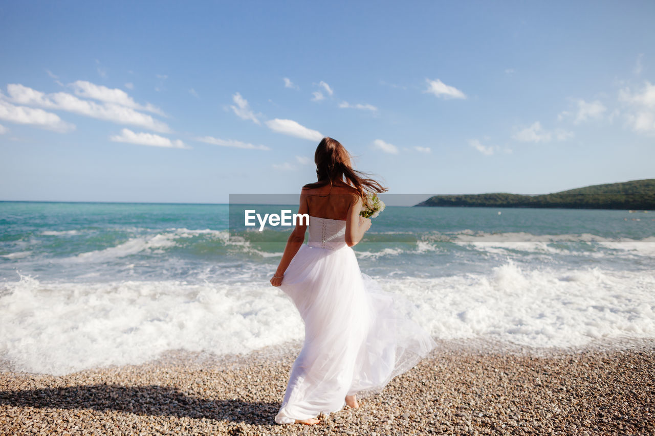 Rear view of bride holding flowers at beach on sunny day