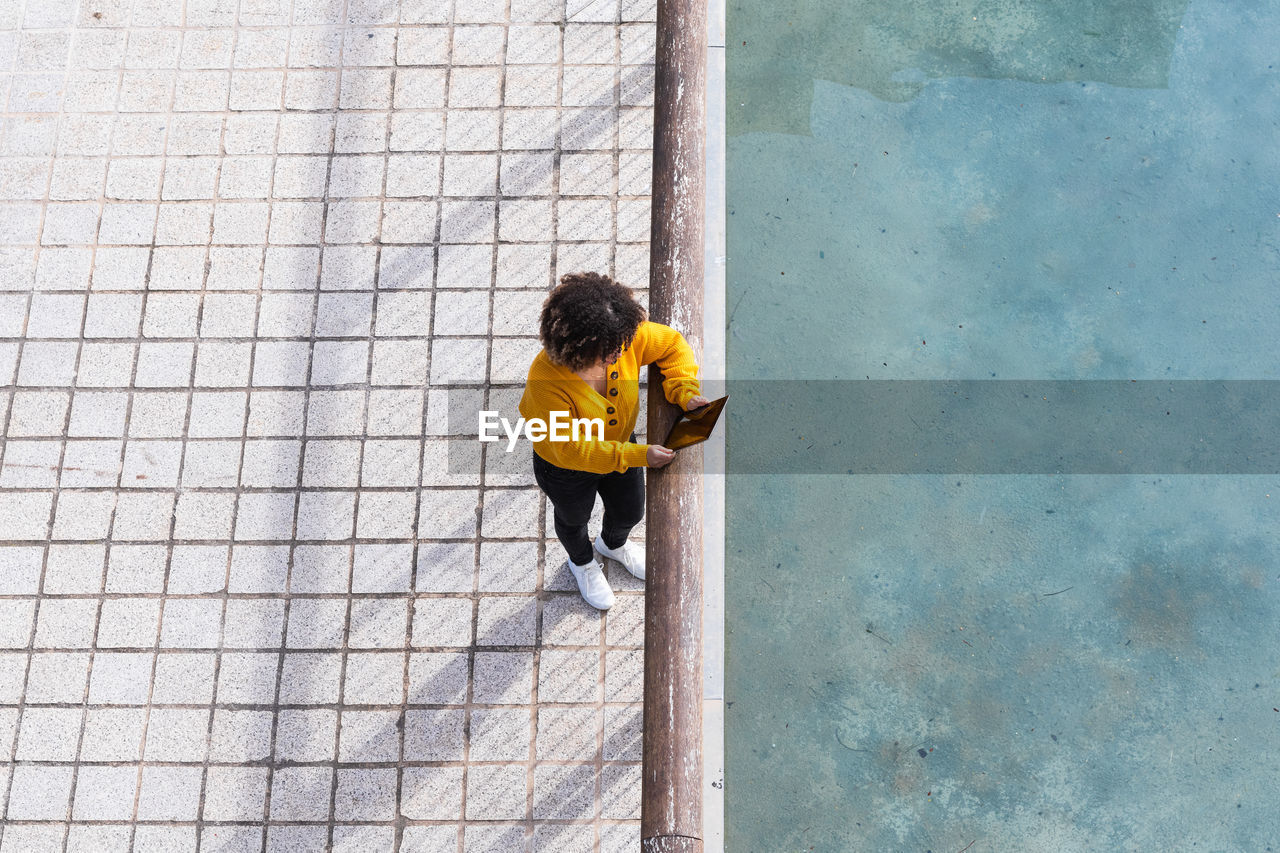 From above remote view of female with afro hairstyle standing near metal railing in urban area of city while using a tablet device