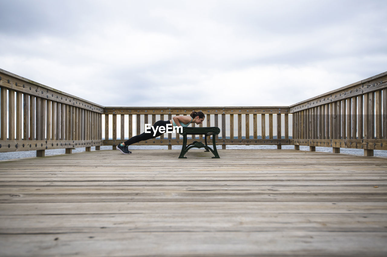 Side view of woman doing push-ups on bench at pier against cloudy sky