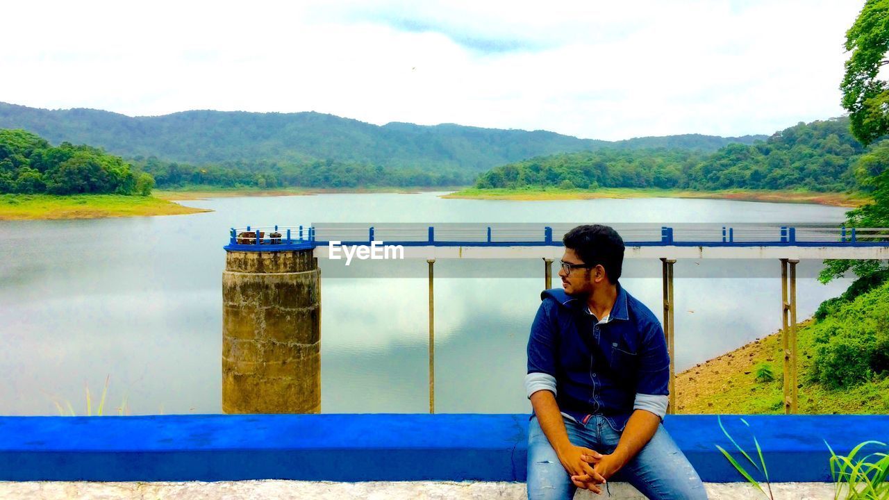 Young man sitting on retaining wall against vazhani dam