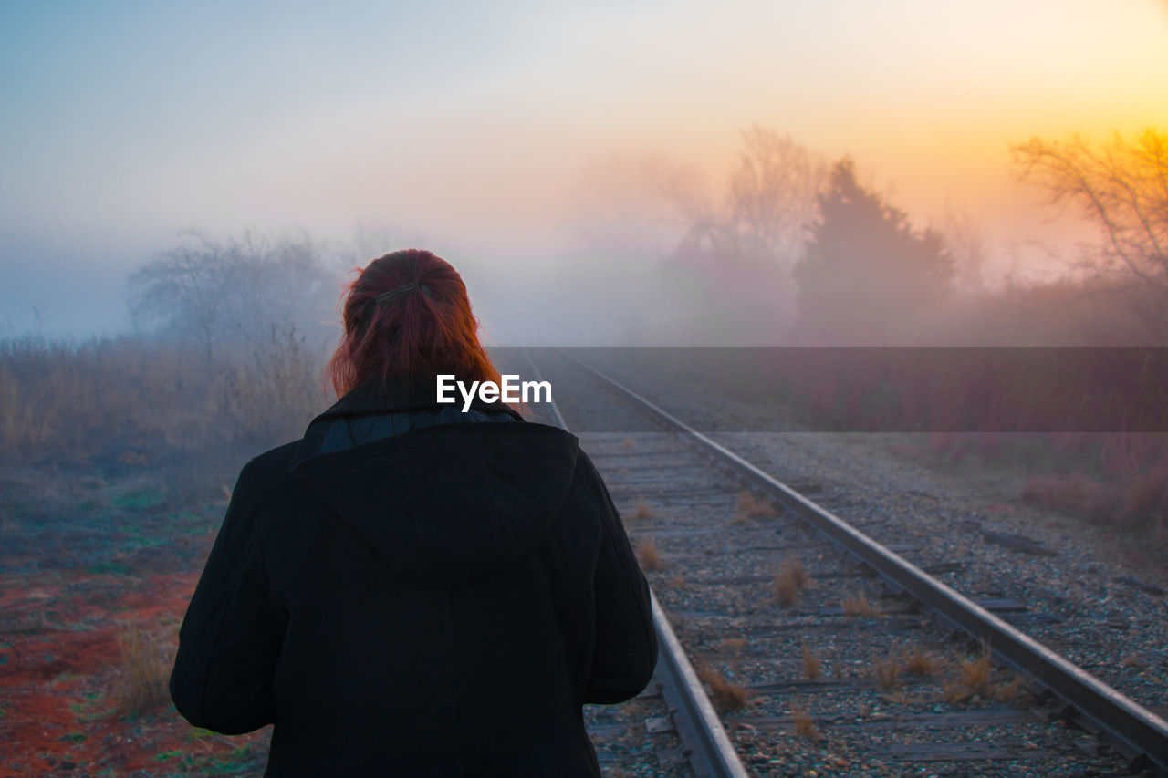 Rear view of person standing by railroad track during sunset