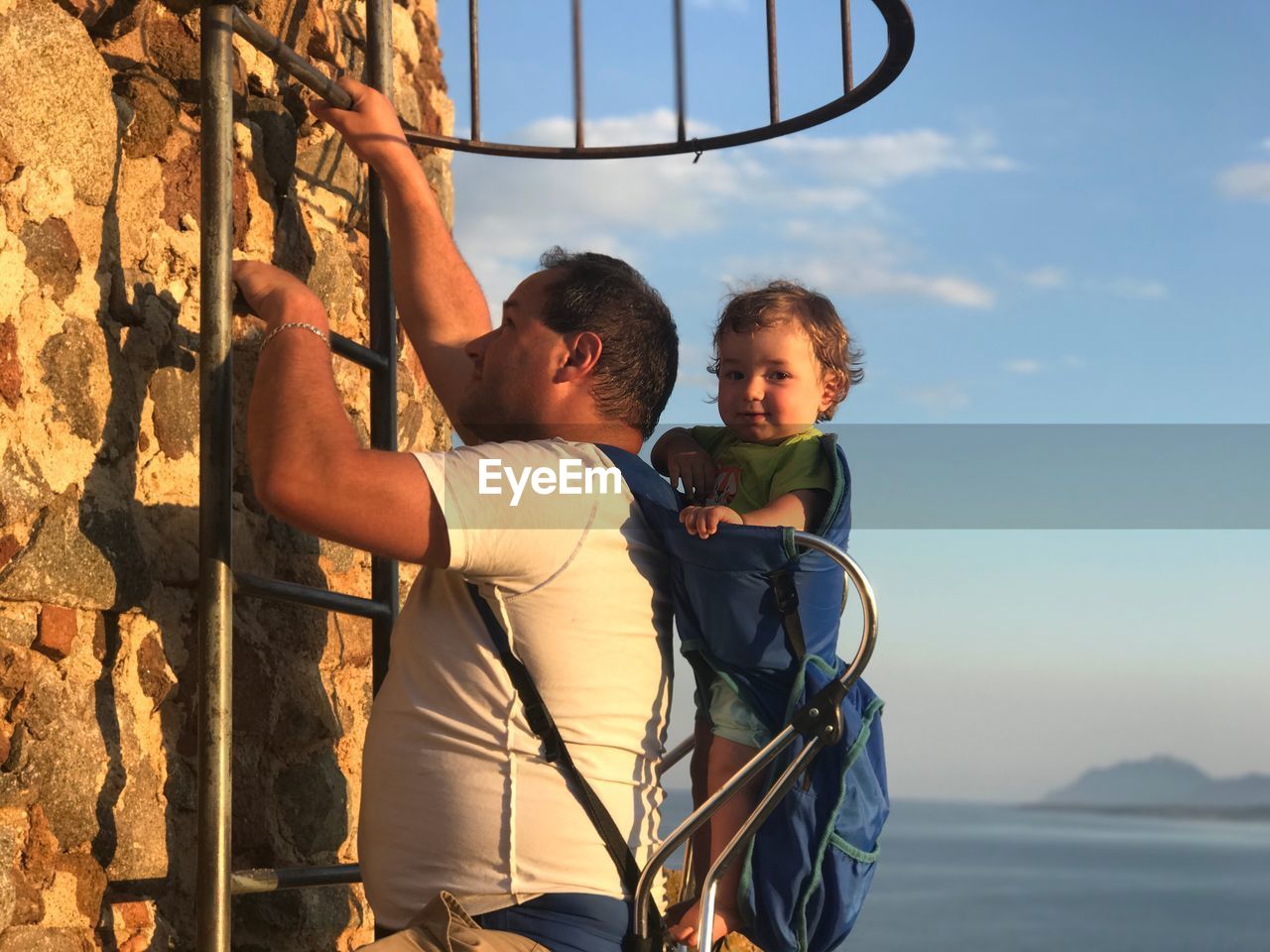 Side view of father climbing ladder while carrying son against sky