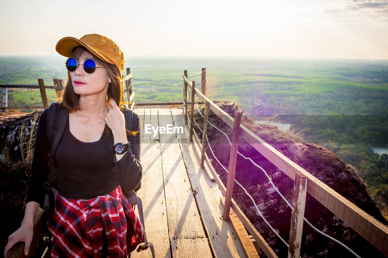 YOUNG WOMAN WEARING SUNGLASSES STANDING ON RAILING AGAINST SKY
