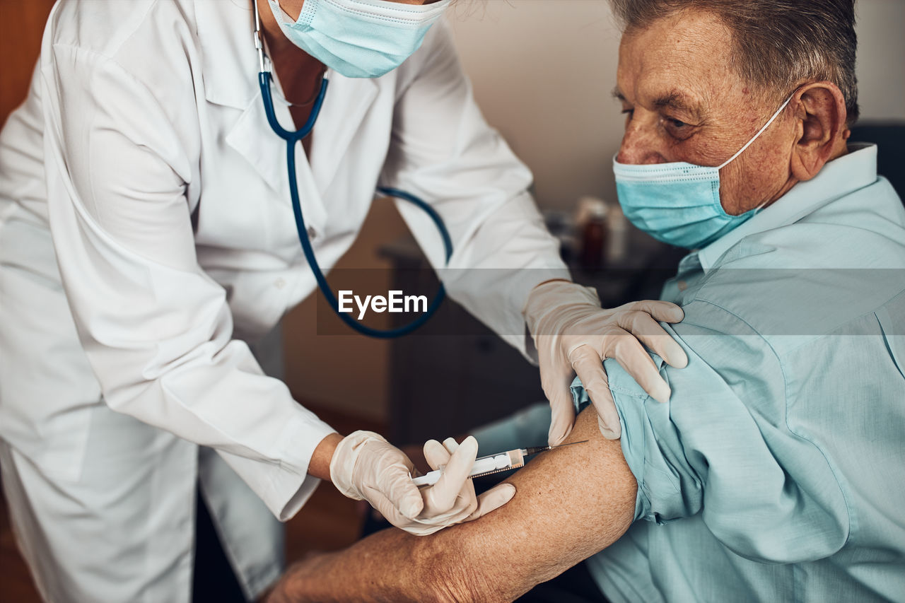 Doctor holding syringe with vaccine and making injection to senior patient with medical mask