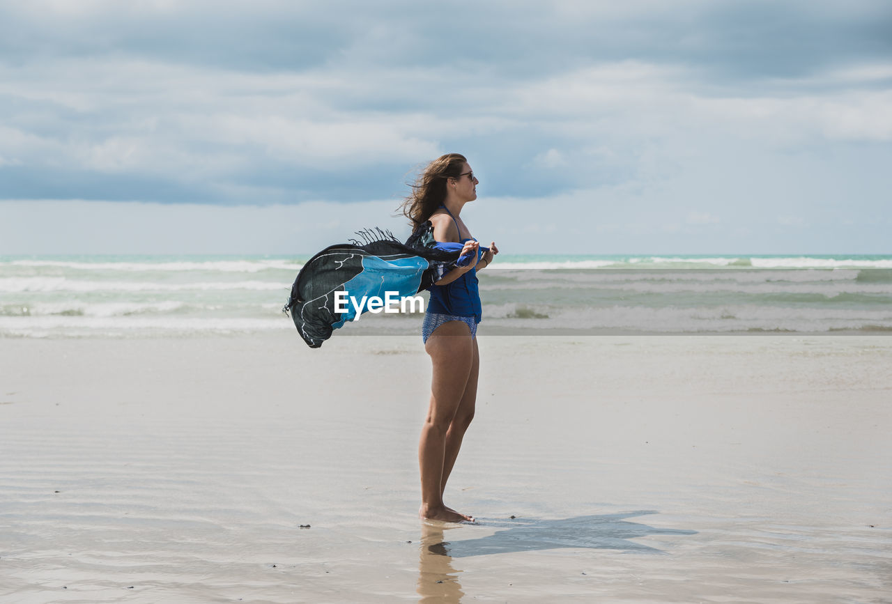 Side view of woman in swimwear holding sarong while standing at beach against cloudy sky during sunny day