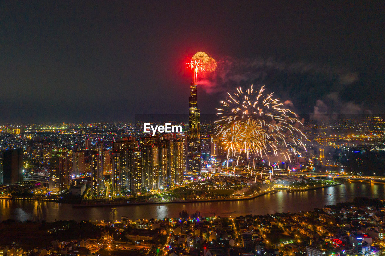 FIREWORK DISPLAY OVER RIVER AGAINST ILLUMINATED BUILDINGS