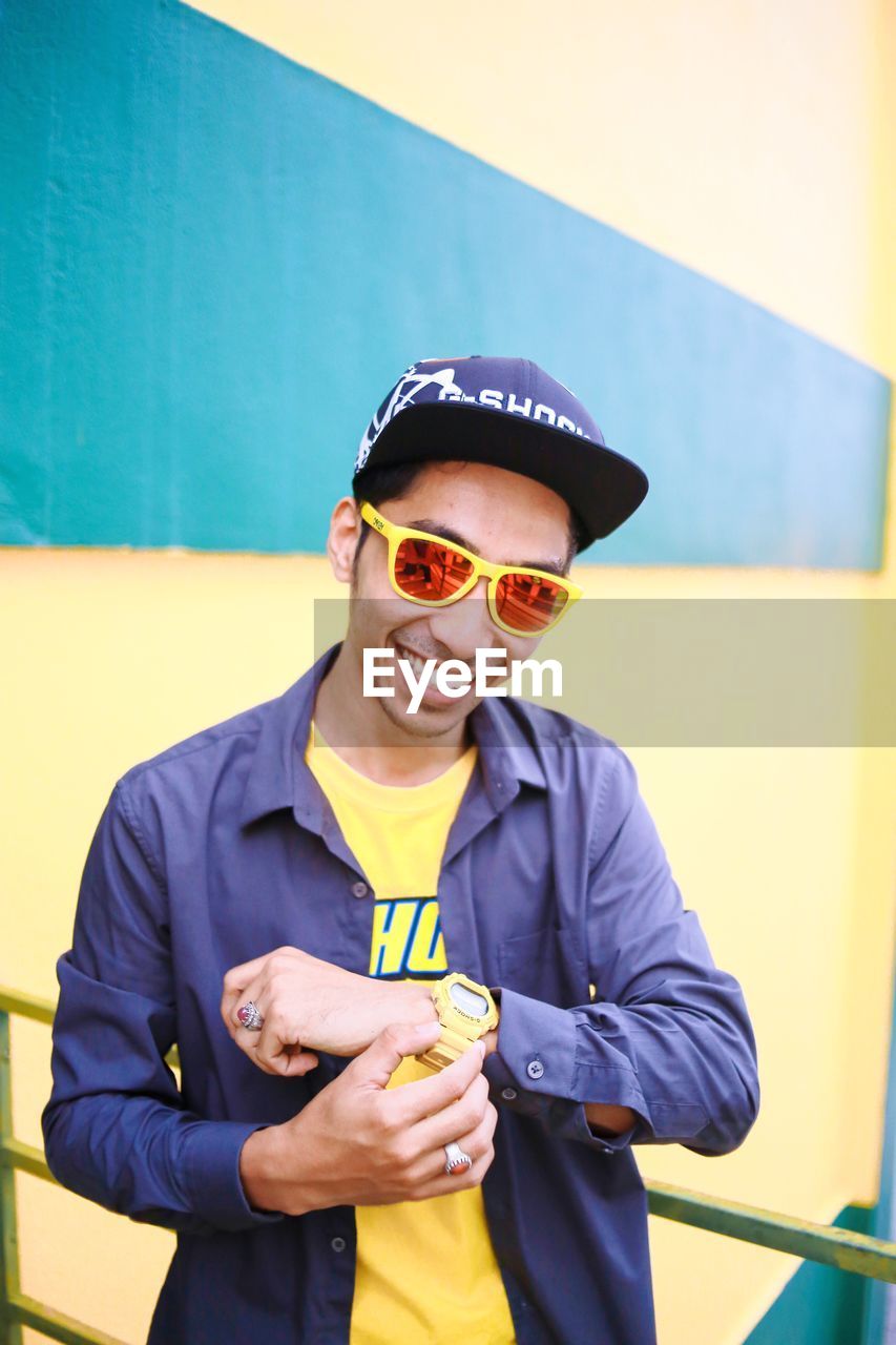 PORTRAIT OF HAPPY MAN WEARING SUNGLASSES STANDING AGAINST WALL