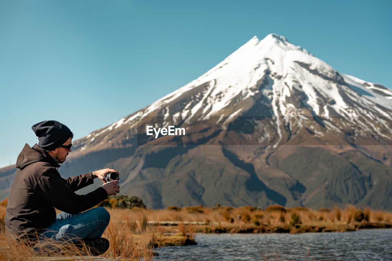 MAN SITTING ON SNOWCAPPED MOUNTAINS AGAINST SKY