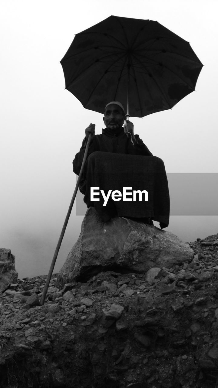 Low angle view of man holding umbrella and cane while sitting on rock against sky