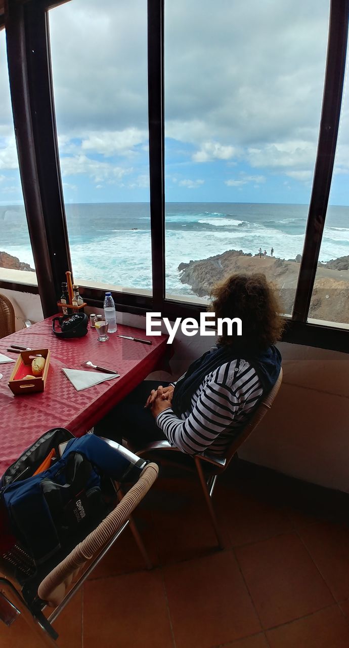 WOMAN SITTING ON TABLE BY SEA AGAINST SKY