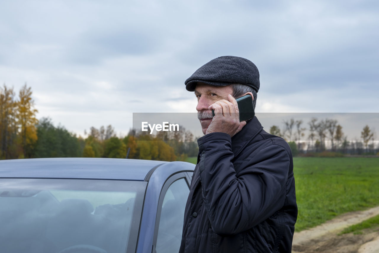 Adult pensioner gets into car and talks on smartphone against backdrop of rural landscape in autumn.