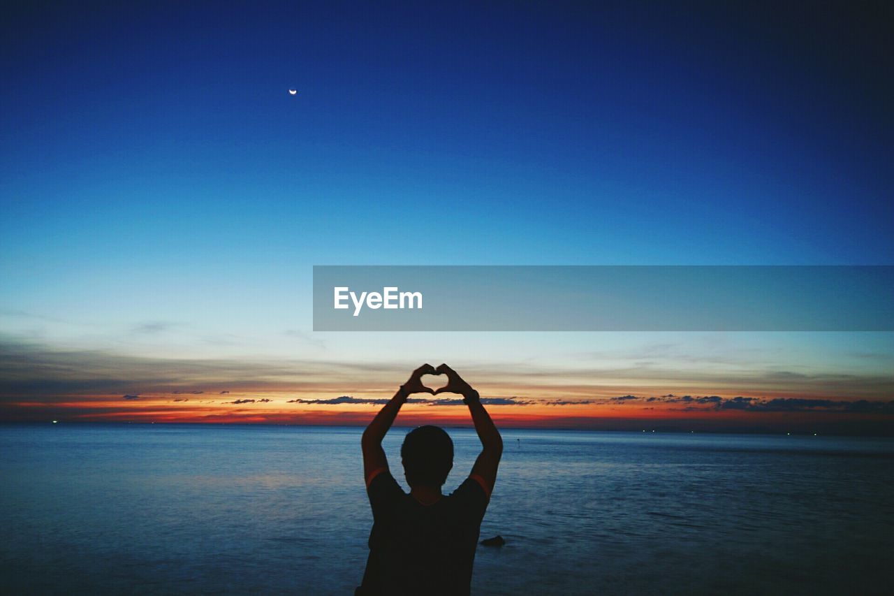 Silhouette man making heart shape while standing at beach against blue sky during sunset