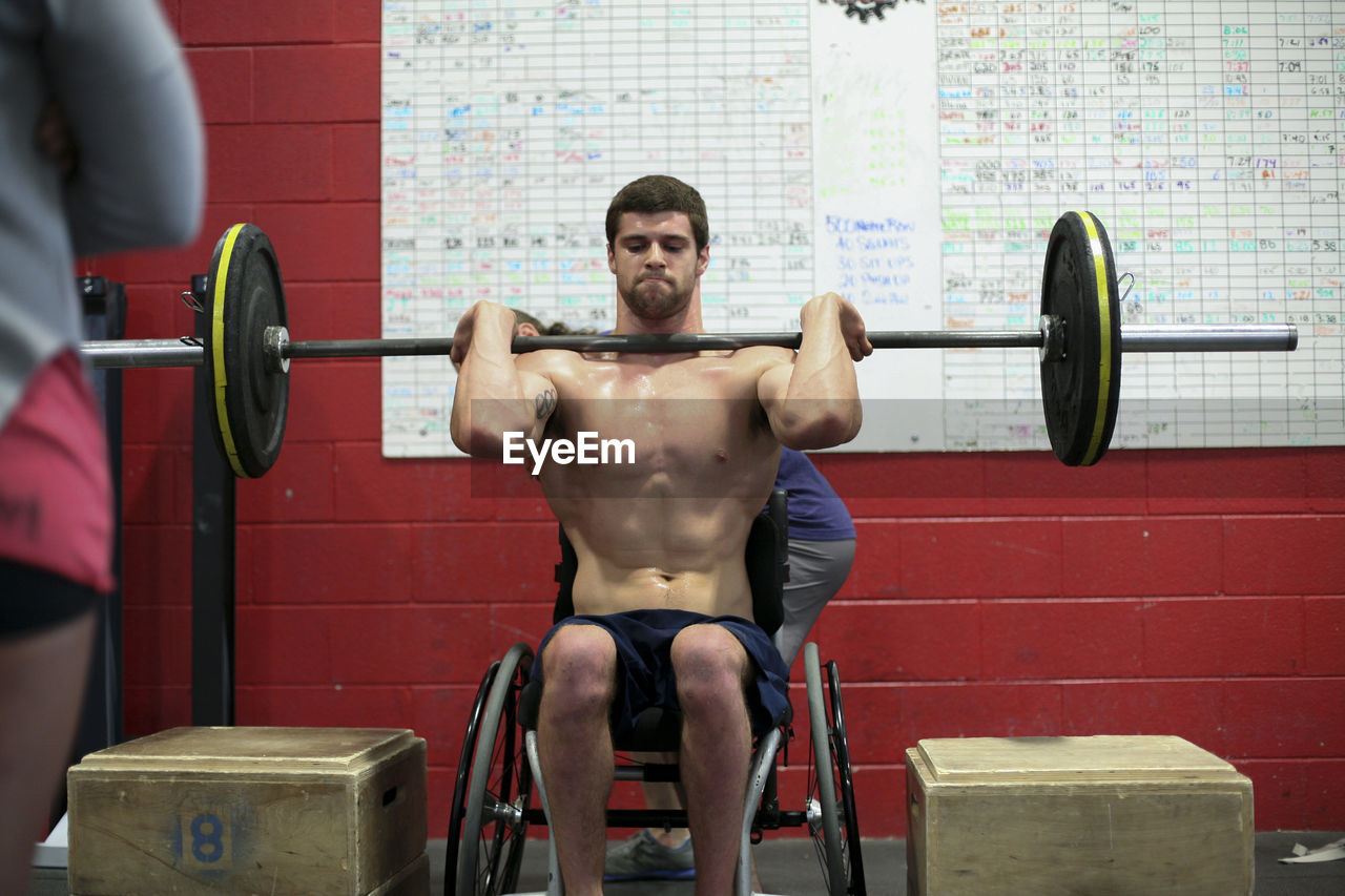 Shirtless male adaptive athlete lifting barbell while sitting on wheelchair against wall in gym