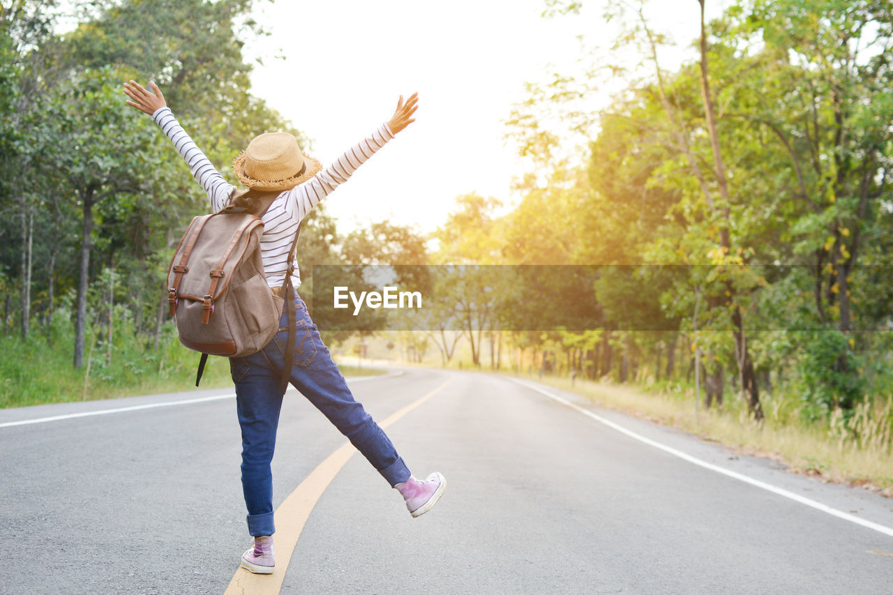 Rear view of playful woman with backpack standing on road amidst trees